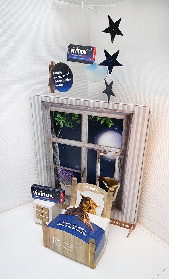 Schaufensterdekoration von pod GmbH für das Medikament vivinox - bestehend aus einem Mobile mit Sternen und einer vivinox-Packung, einem Hintergrundaufsteller mit Blick aus dem Fenster auf den Nachthimmel und einem Bett mit schlafender Eule im Vordergrund