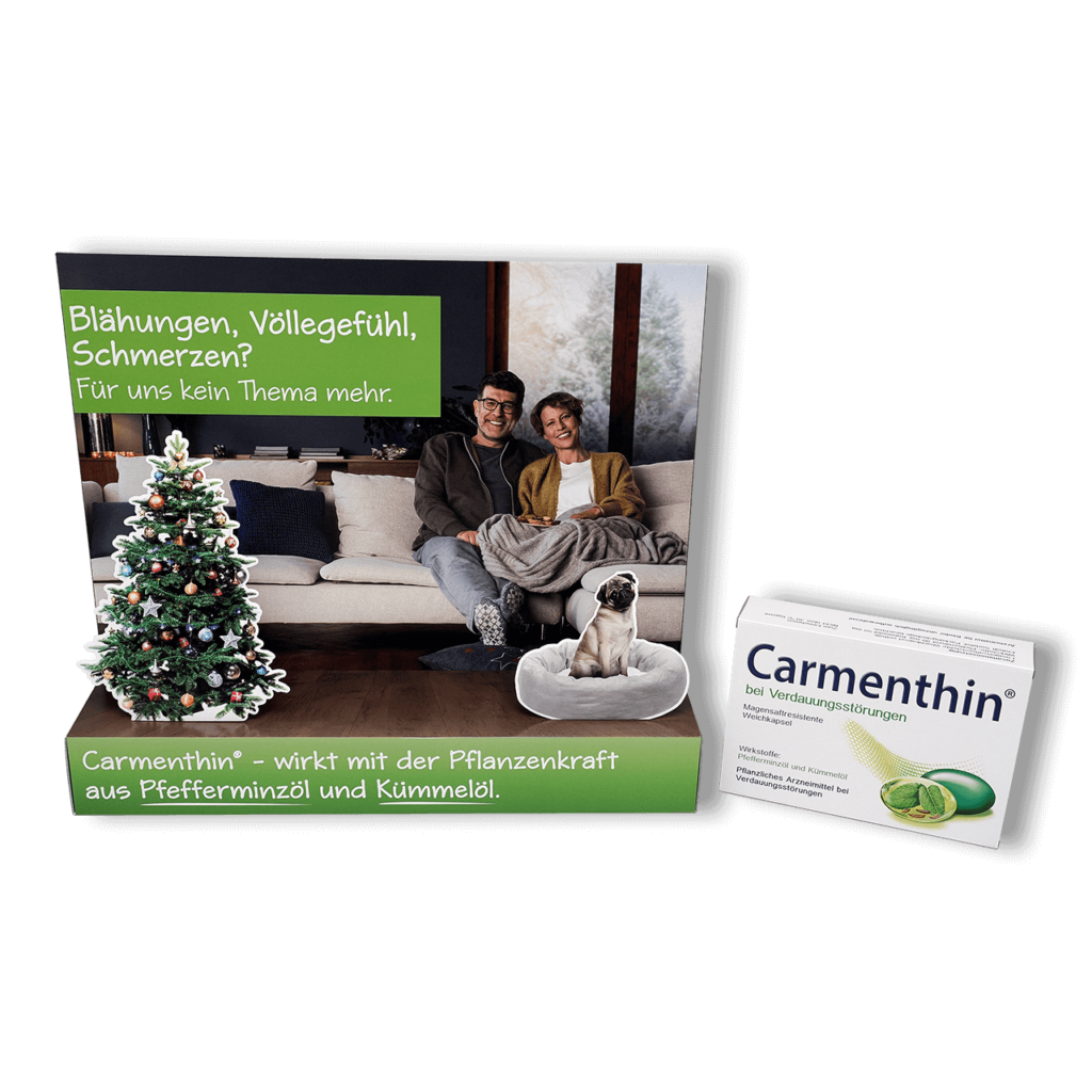 Schaufensterdekoration von pod GmbH für das Medikament Carmethin mit der Abbildung eines lächelnden Paares auf ihrem Sofa im Wohnzimmer, im Vordergrund ein geschmückter Weihnachtsbaum